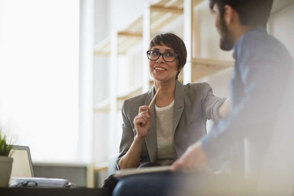 Two cheerful colleagues smiling while discussing something in modern office, focus on stylish businesswoman wearing creative haircut and glasses
