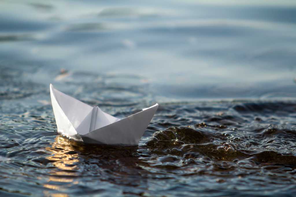 Close-up of simple small white origami paper boat floating in blue clear river or sea water under bright summer sky. Beauty of nature, freedom, dreams and fantasies concept.