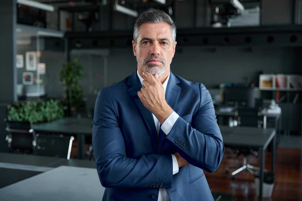 Serious confident mid aged business man, thoughtful doubtful company ceo executive wearing blue suit standing in office holding hand in chin looking at camera thinking, making decision, feeling doubt.