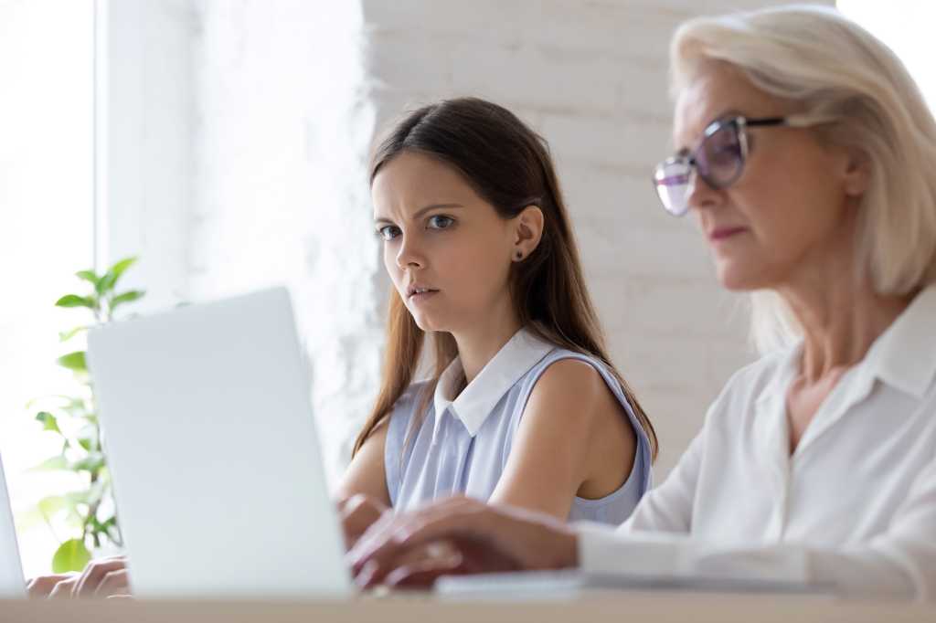 Surprised young woman worker sit at office desk work near coworker feel stunned amazed looking at senior colleague laptop screen, shocked female employee spy peep at workmate computer in office