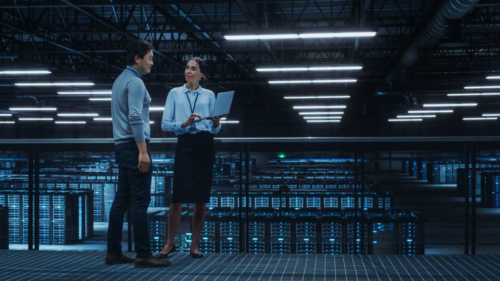 Data Center Female System Administrator and Male IT Specialist Talk, Use Laptop Computer. Information Technology Engineers work on Cyber Security Network Protection in Cloud Computing Server Farm.