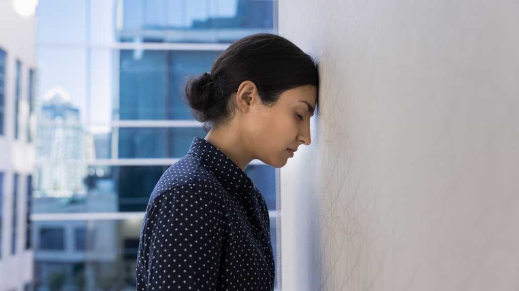 Stressed tired young Indian employee woman banging head against wall in burnout, feeling exhausted, getting professional crisis, problems, bankruptcy. Side view with business building background