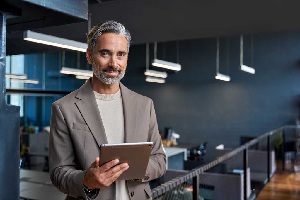 Portrait of confident mature business man executive standing in corporate office using digital tablet. Middle aged professional businessman manager wearing suit posing at work in modern space.