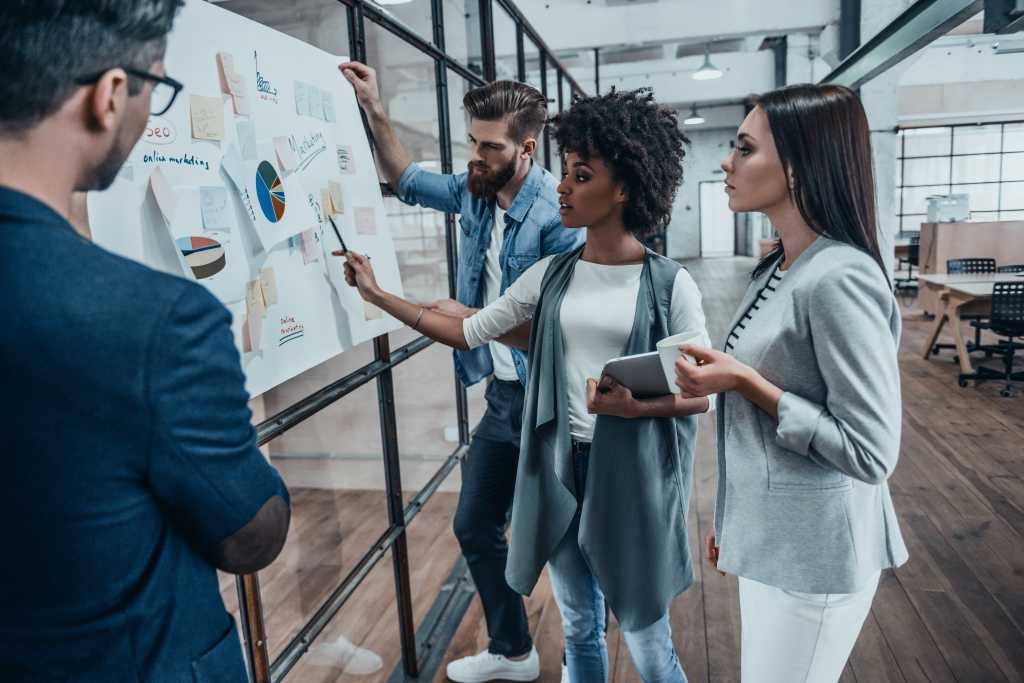 Collaboration is a key to best results. Group of young modern people in smart casual wear planning business strategy while young woman pointing at infographic displayed on the glass wall in the office