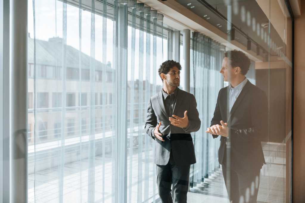 Male business professionals discussing while walking in office corridor seen through glass